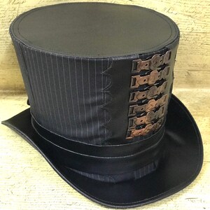 May include: A black top hat with a pinstripe pattern and a band of black leather around the base. The hat has a series of decorative metal buckles on the side.
