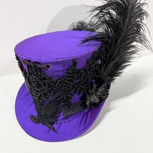 Pu&ograve; includere: Un cappello a cilindro viola con decorazioni nere, tra cui perline e una grande piuma. Il cappello presenta dettagli in pizzo nero e un velo nero trasparente, creando un contrasto sorprendente.