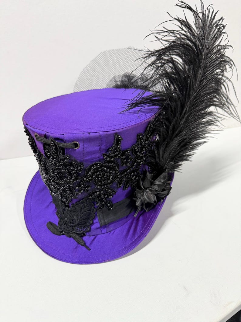 Pu&ograve; includere: Un cappello a cilindro viola con decorazioni nere, tra cui perline e una grande piuma. Il cappello presenta dettagli in pizzo nero e un velo nero trasparente, creando un contrasto sorprendente.