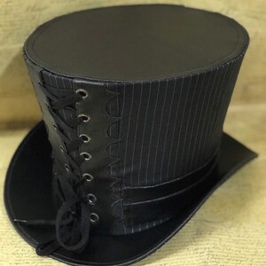 May include: A black top hat with a pinstripe pattern and a black leather band with a lace-up detail. The hat has a wide brim and a tall crown.