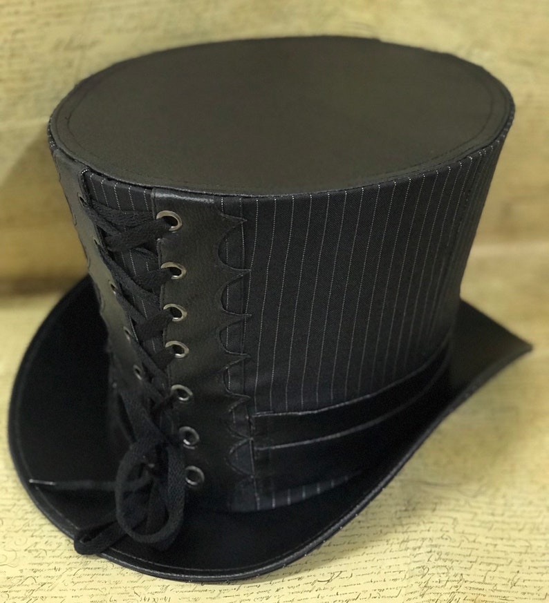 May include: A black top hat with a pinstripe pattern and a black leather band with a lace-up detail. The hat has a wide brim and a tall crown.