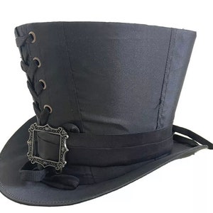 Op de afbeelding: Een zwarte hoge hoed met een brede rand en een hoge, cilindrische kroon. De hoed heeft een decoratieve band rond de basis en een zilverkleurige gesp. De zijkant van de hoed heeft een veterdetail.