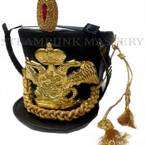 Könnte beinhalten: Schwarzer Militärhut mit goldenen Verzierungen. Er verfügt über ein goldenes Emblem mit Kronen- und Adlerdesign, eine goldene geflochtene Bordüre und eine goldene Quaste. Der Hut hat eine kleine, goldene, ovale Dekoration oben. Der Text "Steampunk Mastery" ist sichtbar.