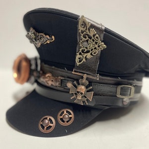 Op de afbeelding: Een zwarte steampunk-hoed met een schedel en gekruiste beenderen ontwerp, een leren band en decoratieve metalen accenten.