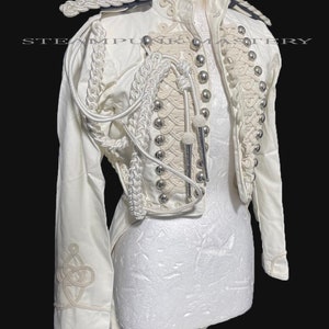 May include: A white military-style jacket with a high collar and intricate white braid detailing. The jacket has a fitted waist and a long tail. The jacket is decorated with silver buttons and has a unique design on the sleeves.