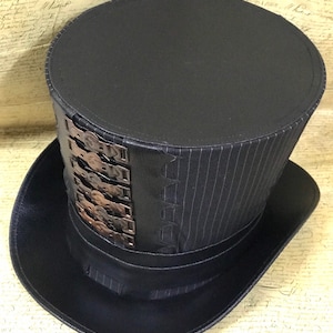 May include: A black top hat with a striped fabric band and a row of metal accents. The hat has a wide brim and a tall, cylindrical crown.