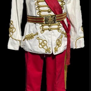 May include: A white military-style jacket with gold trim and buttons, a red sash, and red pants. The jacket has a gold embroidered design and a gold belt with a buckle. The pants have a gold stripe down the side.