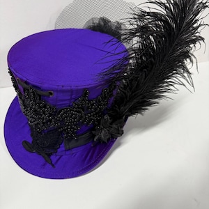 Pu&ograve; includere: Un cappello a cilindro viola con abbellimenti neri e una grande piuma nera. Il cappello presenta un design in pizzo nero alla base e un velo nero. Un accessorio unico.