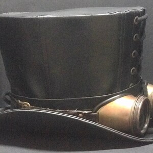 Puede incluir: Un sombrero de copa de cuero negro con un accesorio de gafas steampunk de lat&oacute;n. El sombrero tiene una parte posterior con cordones y un ala ancha.