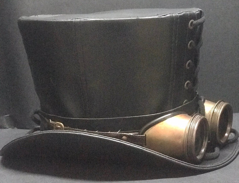 Puede incluir: Un sombrero de copa de cuero negro con un accesorio de gafas steampunk de lat&oacute;n. El sombrero tiene una parte posterior con cordones y un ala ancha.