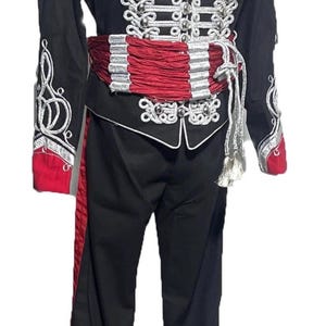 Könnte beinhalten: Eine schwarze und rote Militärjacke mit silbernen Verzierungen und Knöpfen. Die Jacke hat eine rote Schärpe und silberne Stickereien an den Ärmeln. Die Jacke wird mit einer schwarzen Hose mit einem roten Streifen an der Seite kombiniert.