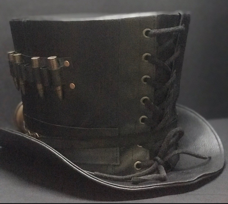 Puede incluir: Un sombrero de copa de cuero negro con un detalle de encaje en el lateral y una banda de balas alrededor de la base.