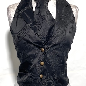 May include: Black halter vest with a matching neck tie. The vest features a subtle, ornate pattern and is fastened with three gold-colored buttons. The vest is displayed on a mannequin.