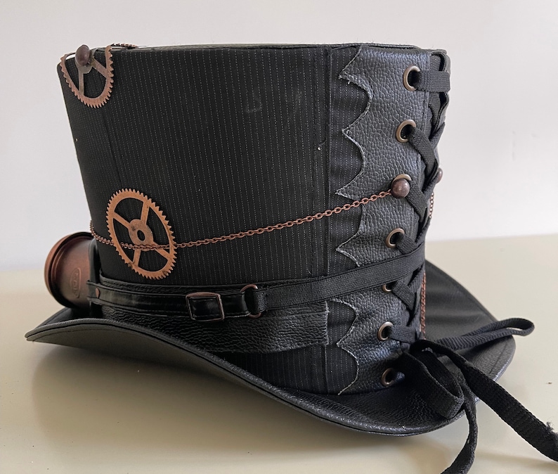 Pu&ograve; includere: Un cappello a cilindro steampunk nero con ingranaggi color rame e una piccola lente rotonda. Il cappello presenta una fascia in pelle nera con fibbia, un design allacciato e una catena decorativa. Il cappello &egrave; realizzato in tessuto a righe.