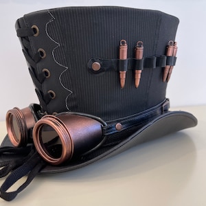 May include: A black top hat with a black and brown band, a black and brown lace-up detail, and a row of copper-colored bullets. The hat is paired with a pair of black and copper-colored goggles.