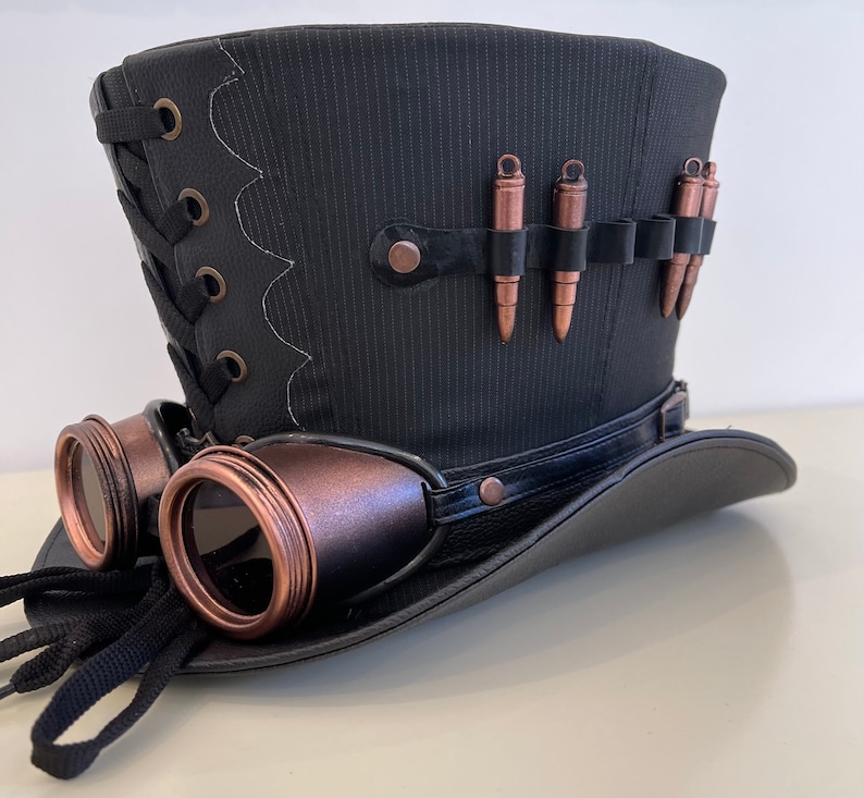 May include: A black top hat with a black and brown band, a black and brown lace-up detail, and a row of copper-colored bullets. The hat is paired with a pair of black and copper-colored goggles.