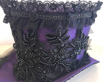 May include: A purple top hat with black lace and bead embellishments. The hat has a wide brim and a tall crown.