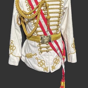 May include: White military-style jacket with gold embroidery, buttons, and epaulets. A red and white sash crosses the front, complemented by a gold belt and tassel. The text "STEAMPUNK MASTERY" is visible.