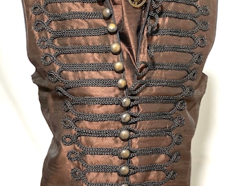 Puede incluir: Un chaleco marrón sin mangas con ribetes trenzados negros y botones dorados. El chaleco tiene un cuello alto y una punta en la parte delantera.