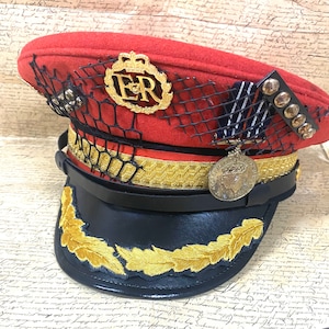 Può includere: Un cappello in stile militare rosso e nero con finiture dorate e un emblema di corona dorata con le lettere "ER" su di esso. Il cappello ha una fascia in pelle nera con una fibbia dorata e una rete a maglia nera sulla parte superiore.