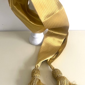 May include: A gold satin sash with two tassels. The sash is striped and has a shiny finish.