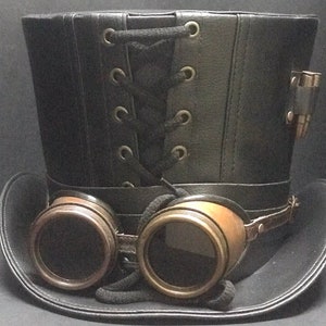 Puede incluir: Un sombrero de copa de cuero negro con un detalle de encaje y un par de gafas de lat&oacute;n unidas al borde.