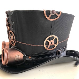 Pu&ograve; includere: Un cappello a cilindro nero con ingranaggi e occhiali color rame, che evoca un'estetica steampunk. Il cappello presenta un tessuto strutturato, un bordo simile alla pelle e dettagli a catena decorativi. Gli occhiali hanno il testo "SOL".
