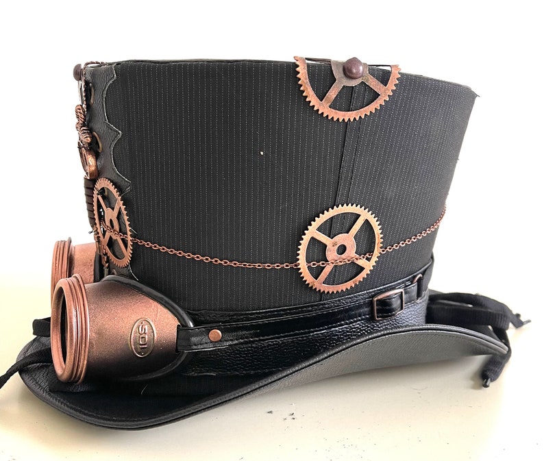 Pu&ograve; includere: Un cappello a cilindro nero con ingranaggi e occhiali color rame, che evoca un'estetica steampunk. Il cappello presenta un tessuto strutturato, un bordo simile alla pelle e dettagli a catena decorativi. Gli occhiali hanno il testo "SOL".