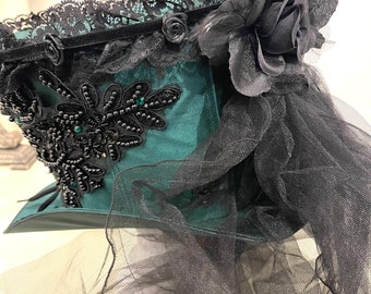 May include: A dark green top hat with black lace trim, a black velvet ribbon, and a black rose with a feather. The hat is adorned with black beaded embellishments and a black tulle veil.
