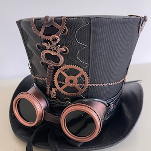 Pu&ograve; includere: Cappello a cilindro steampunk nero con ingranaggi color rame, una chiave e occhiali protettivi attaccati. Il cappello ha un aspetto in pelle strutturata con un pannello in tessuto a righe. Un nastro nero &egrave; attaccato agli occhiali.