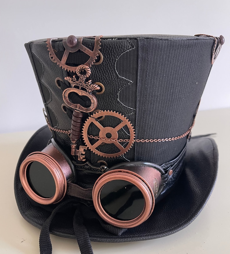 Pu&ograve; includere: Cappello a cilindro steampunk nero con ingranaggi color rame, una chiave e occhiali protettivi attaccati. Il cappello ha un aspetto in pelle strutturata con un pannello in tessuto a righe. Un nastro nero &egrave; attaccato agli occhiali.