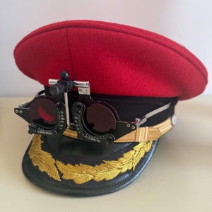 Może przedstawiać: Czerwono-czarna czapka w stylu wojskowym ze złotym haftem i czarną ramką z soczewkami. Soczewki są ciemnoczerwone. Czapka ma daszek i złoty pasek wokół podstawy.
