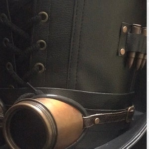 Puede incluir: Un sombrero de copa de cuero negro con una banda de cuero marr&oacute;n y gafas de lat&oacute;n. El sombrero tiene un detalle de encaje y una fila de balas de lat&oacute;n en el lateral.