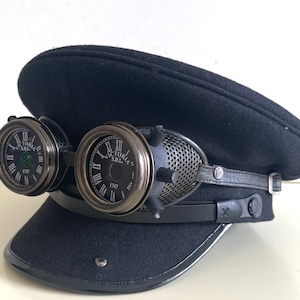 Può includere: Un cappello di feltro nero con una fascia in pelle nera e due occhiali in stile steampunk con quadranti dell'orologio che riportano "Victoria S.D.L. 1747".
