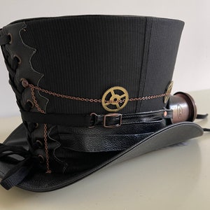 Pu&ograve; includere: Cappello a cilindro nero con dettagli in stile steampunk. Presenta una tesa e una fascia in pelle nera, ingranaggi decorativi e un piccolo oculare color rame. Il cappello ha un motivo a righe verticali e dettagli di allacciatura laterali.