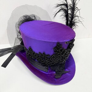 Pu&ograve; includere: Un cappello a cilindro viola con abbellimenti neri, un velo nero e piume nere. Il cappello presenta una finitura satinata e perline nere decorative intorno all'orlo. Un nastro nero &egrave; legato attorno alla base del cappello.