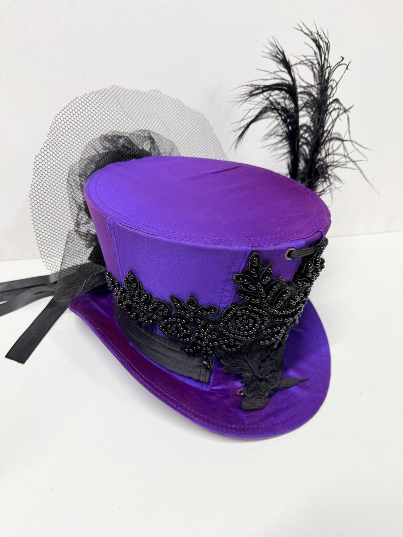 Pu&ograve; includere: Un cappello a cilindro viola con abbellimenti neri, un velo nero e piume nere. Il cappello presenta una finitura satinata e perline nere decorative intorno all'orlo. Un nastro nero &egrave; legato attorno alla base del cappello.