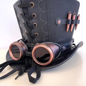 May include: A black top hat with a black leather band and a black lace-up detail on the back. The hat has a pair of copper-colored goggles attached to the front.