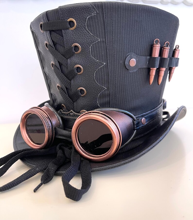 May include: A black top hat with a black leather band and a black lace-up detail on the back. The hat has a pair of copper-colored goggles attached to the front.