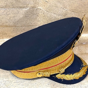 May include: A dark blue military style hat with gold trim and a gold braid around the crown. The hat has a gold leaf design on the side.