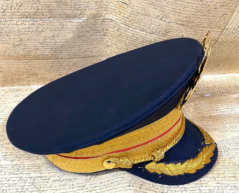 May include: A dark blue military style hat with gold trim and a gold braid around the crown. The hat has a gold leaf design on the side.