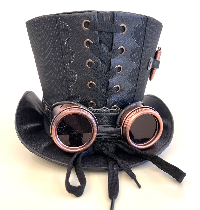 May include: A black top hat with a black lace-up detail and a pair of copper-colored goggles attached to the front. The hat has a black band around the base and a black ribbon tied at the front.