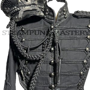 Può includere: Giacca e cappello neri in stile militare con dettagli a teschio. La giacca presenta trecce ornate, bottoni argentati e una fibbia decorativa con un motivo a teschio. Il cappello ha un'emblema a teschio. Il testo "STEAMPUNK MASTERY" è visibile.