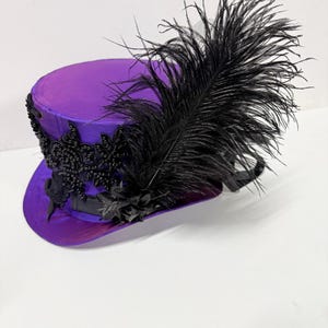 Pu&ograve; includere: Un cappello a cilindro viola con abbellimenti neri e una grande piuma nera. Il cappello presenta una fascia decorativa con perline nere e un nastro di raso nero. Il cappello &egrave; di un vibrante colore viola.