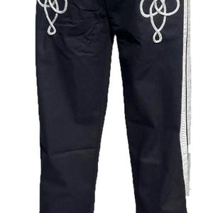 Può includere: Pantaloni neri con dettagli in corda bianca sui lati e lungo le gambe. I dettagli in corda sono in un motivo decorativo. I pantaloni hanno una chiusura a bottone in vita.