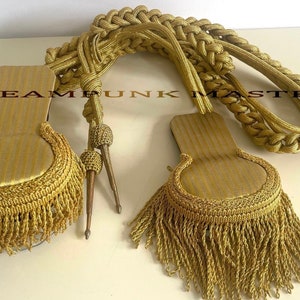 Puede incluir: Un juego de cordones de hombro militares dorados con borlas doradas. Los cordones están trenzados y tienen un nudo decorativo en el extremo. Las borlas están hechas de flecos dorados.