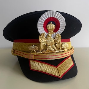 Może przedstawiać: Czarny kapelusz w stylu wojskowym z złotym orłem i koroną, czerwonymi i złotymi ozdobami oraz okrągłą naszywką w kolorze białym, czerwonym i niebieskim.