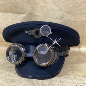 Può includere: Un cappello da capitano in feltro nero con una fascia in pelle e due occhiali rotondi. Il cappello ha un design decorativo a forma di buco della serratura in metallo e una piccola lente d'ingrandimento attaccata alla fascia.