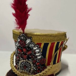 May include: A red, white, and blue striped hat with gold trim and a red feather. The hat is decorated with red and clear rhinestones.