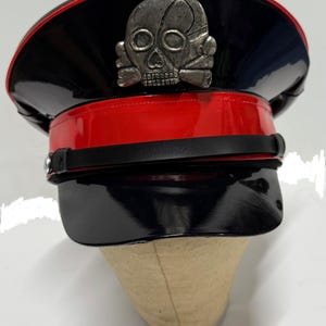 Może przedstawiać: Czarny i czerwony kapelusz w stylu wojskowym z błyszczącym wykończeniem. Kapelusz posiada srebrny emblemat czaszki i czarny pasek. Rondo jest czarne, a opaska czerwona. Kapelusz jest prezentowany na beżowej głowie manekina.